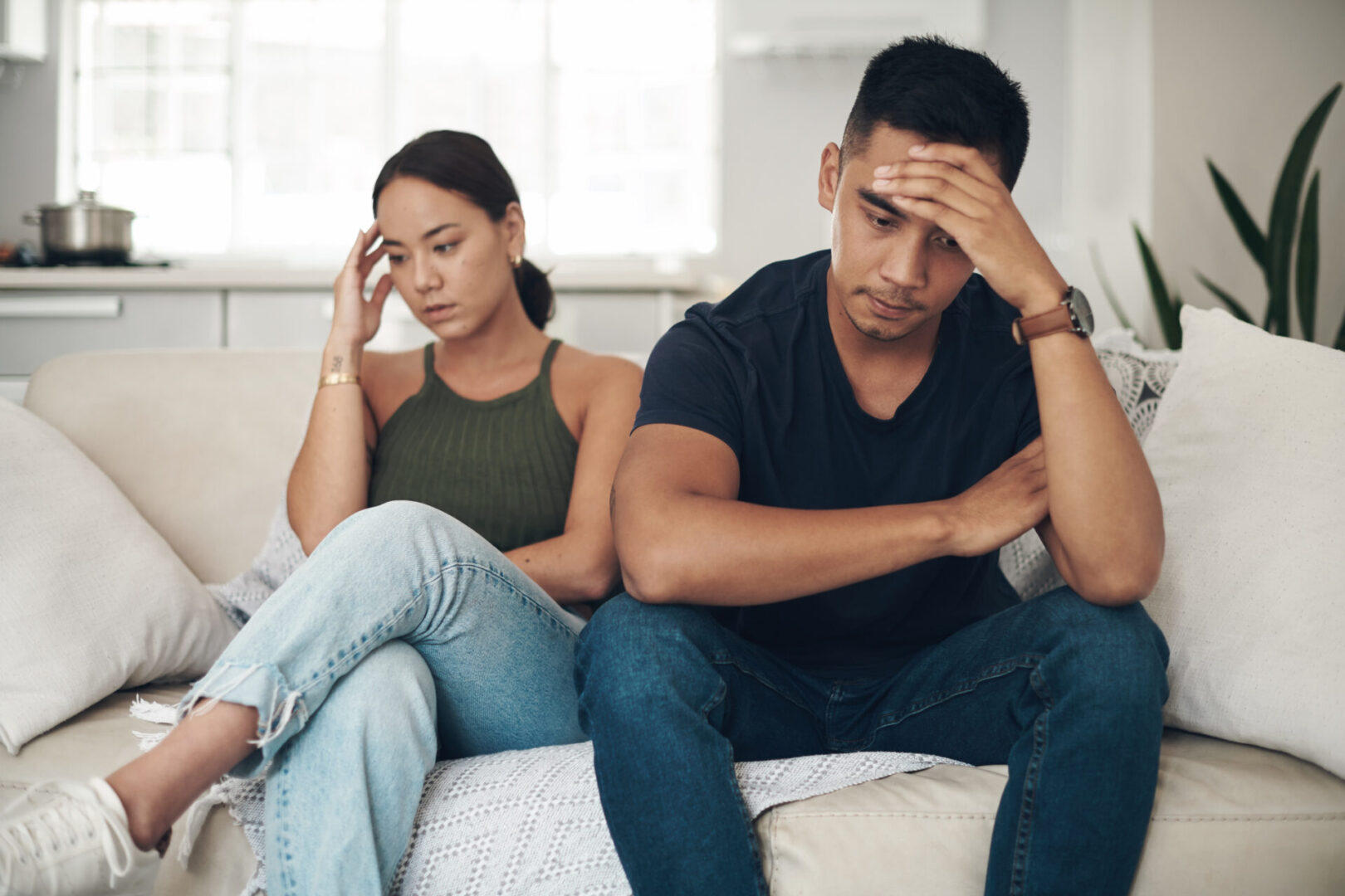 A man and woman sit on a couch, both looking pensive with hands on their foreheads.