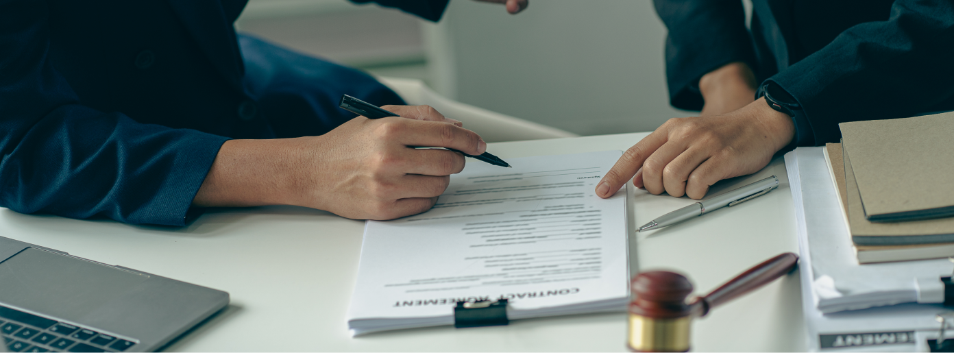 Two people at a desk, one signing a contract. A gavel, a laptop, and documents are nearby.