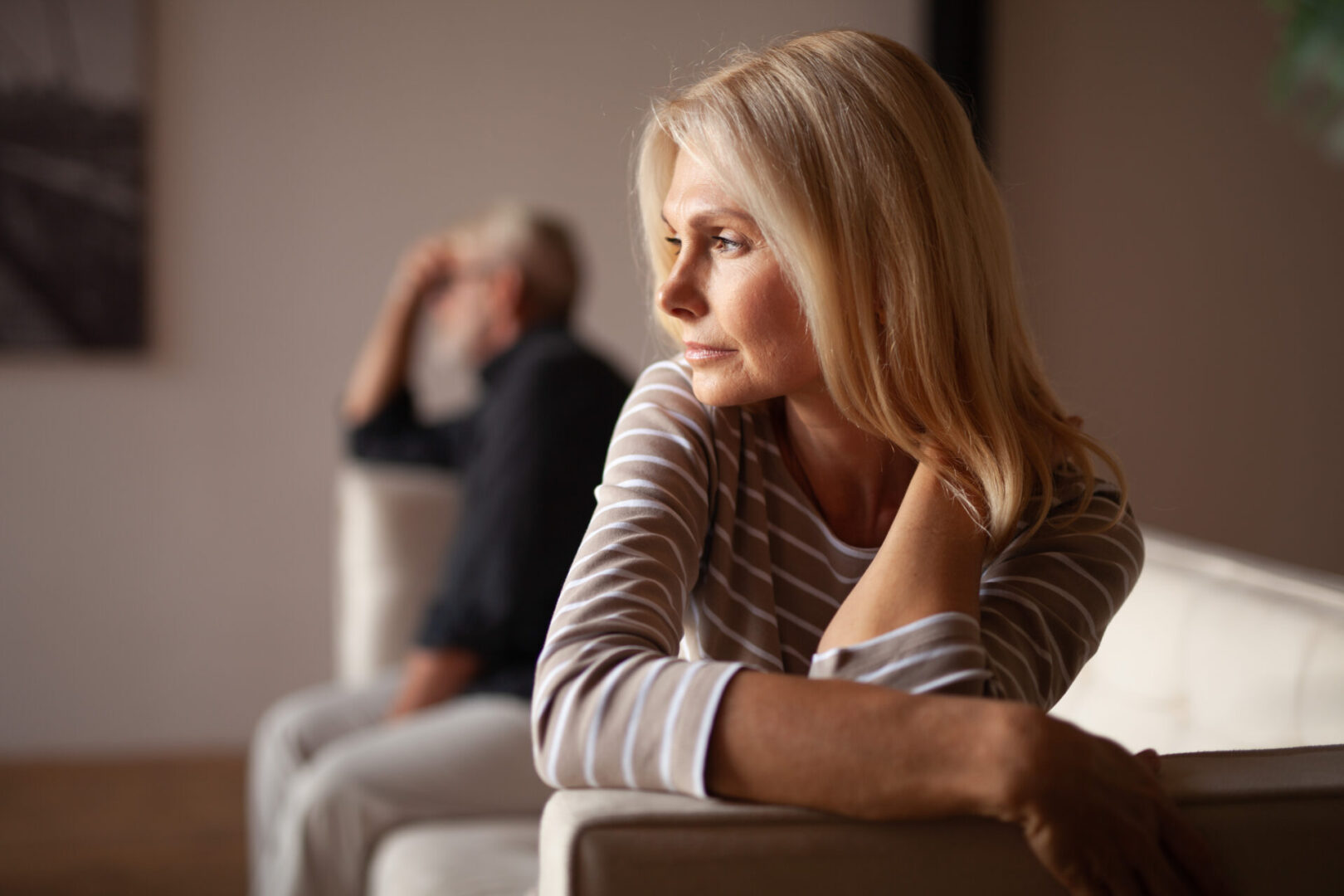 A woman with blonde hair sits on a couch, looking away thoughtfully. In the background, a man sits with his head resting on his hand. The mood appears contemplative.