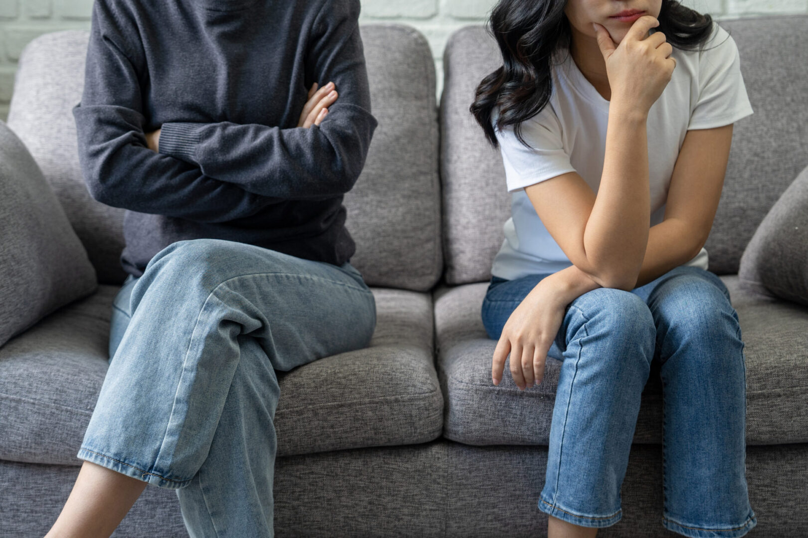 Two people sitting on a gray sofa, both in blue jeans; one wears a gray sweater with arms crossed, and the other in a white t-shirt is resting their chin on their hand.