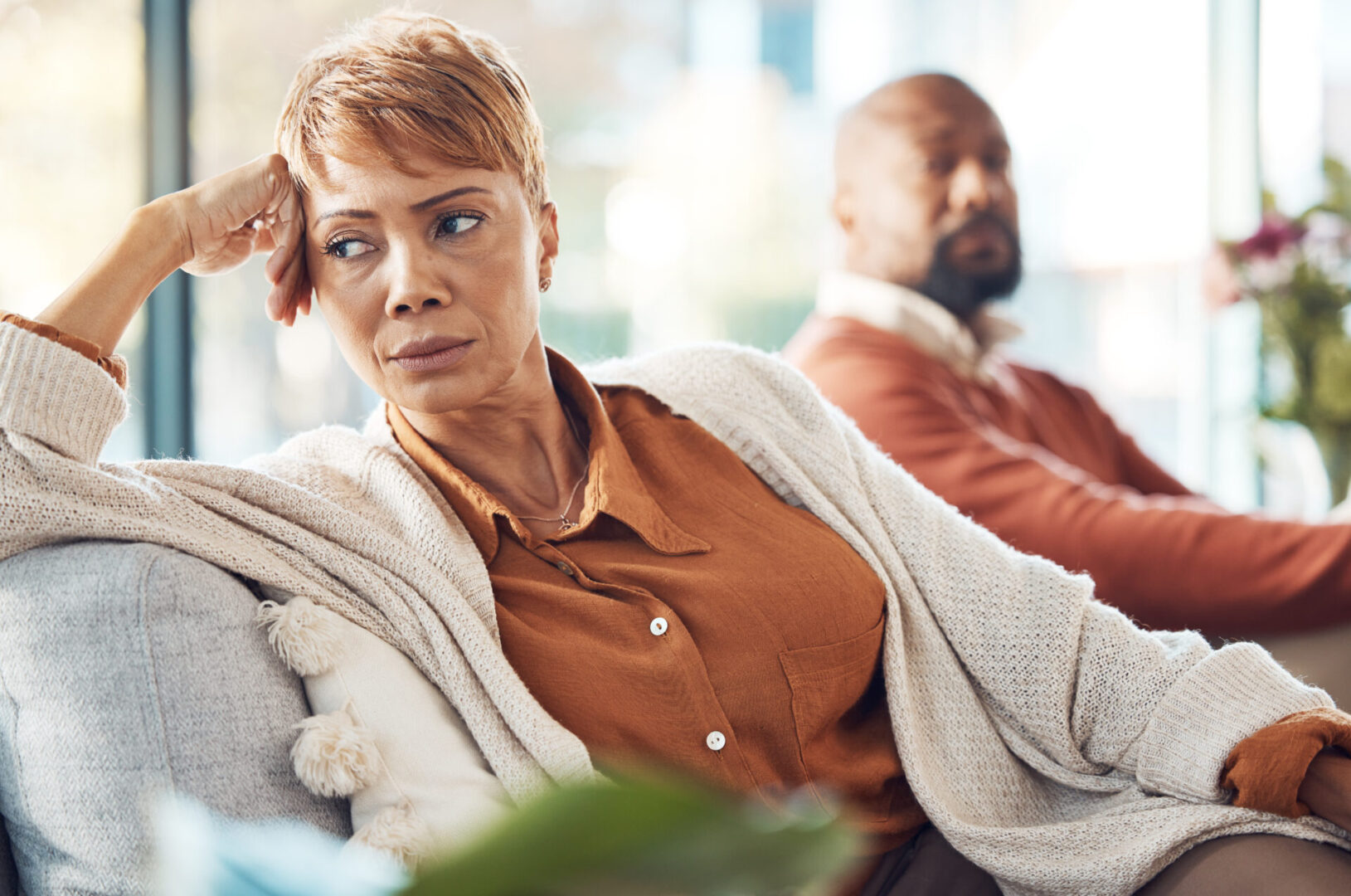 A woman and man sit on a sofa, both appearing thoughtful and distant. The woman looks to the side, resting her head on her hand, while the man sits behind her, slightly out of focus.