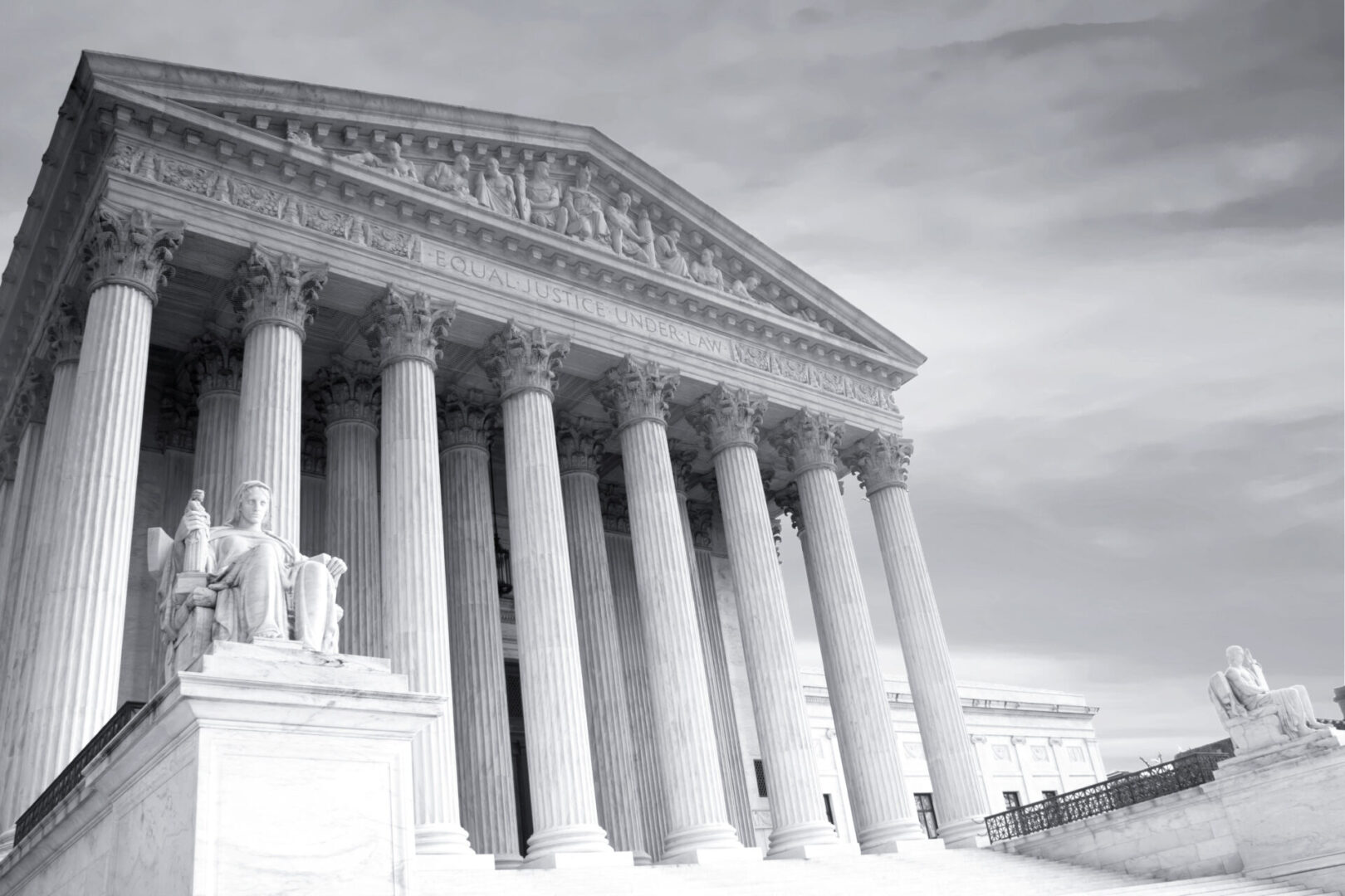 A grayscale photo of a neoclassical building with tall columns and a pediment, featuring two statues at the entrance steps.