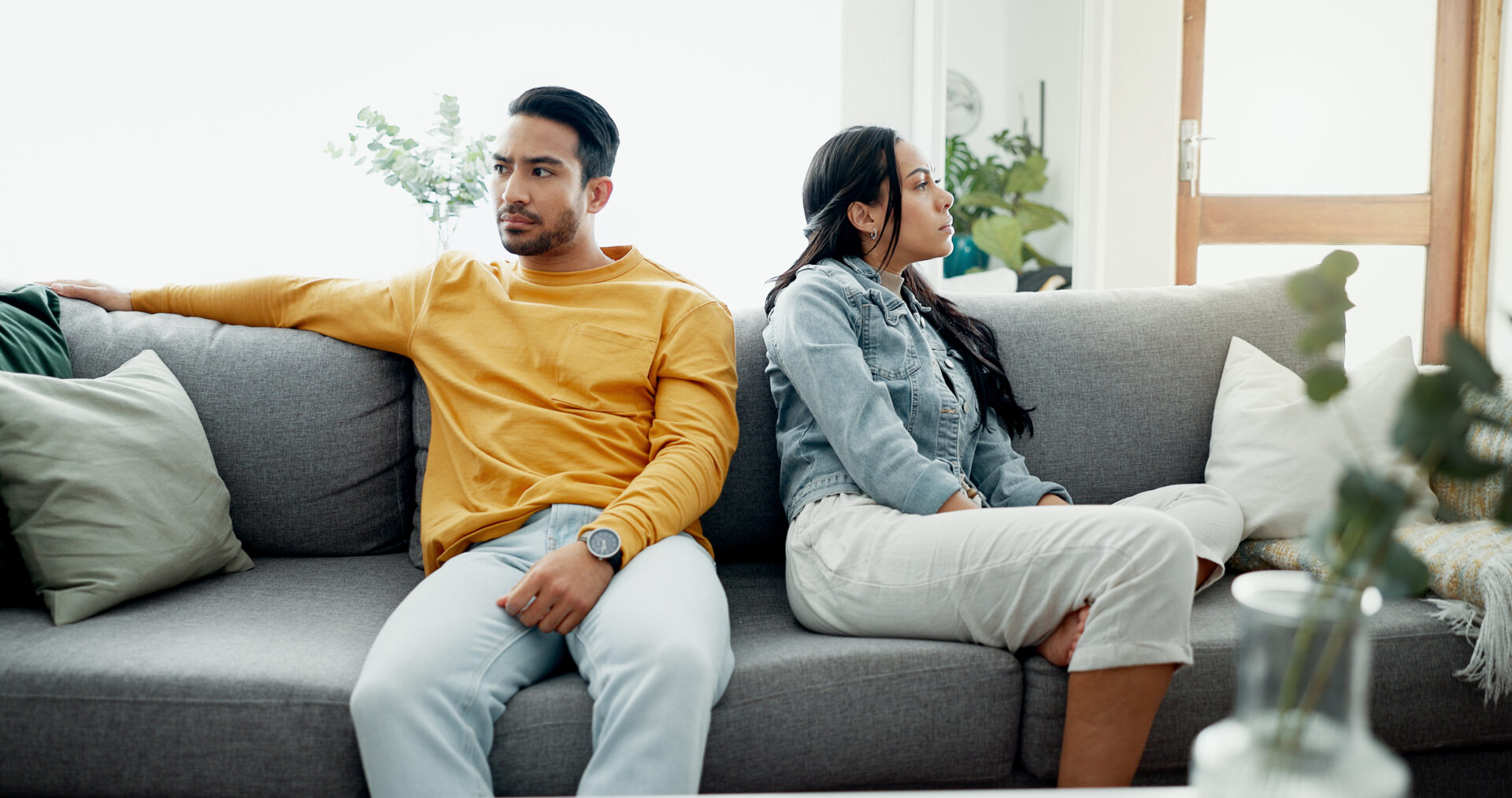 A couple sits on a gray couch, both looking away from each other with serious expressions.