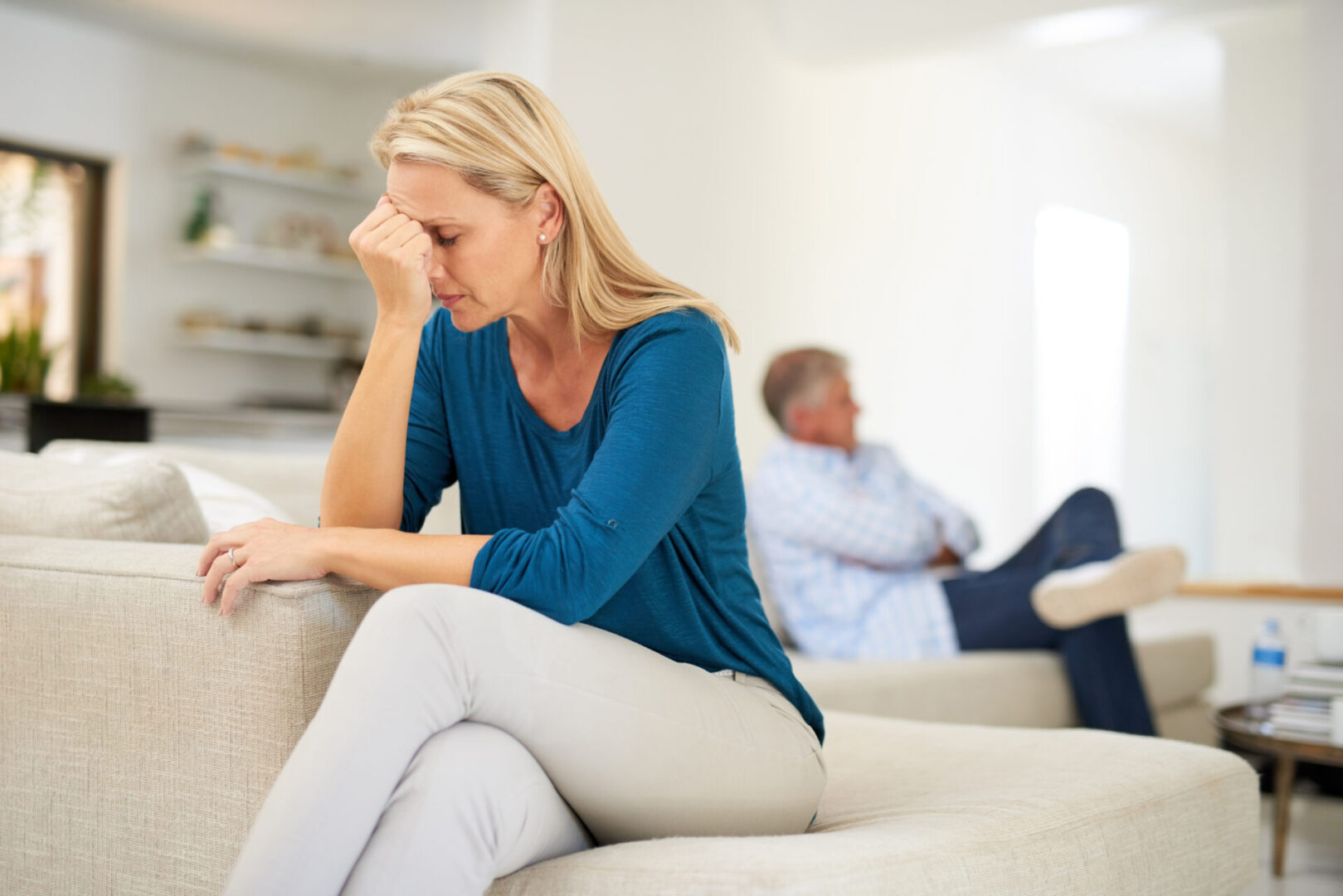 Woman sitting on a couch looking distressed, with an older man in the background sitting with his arms crossed, looking away.