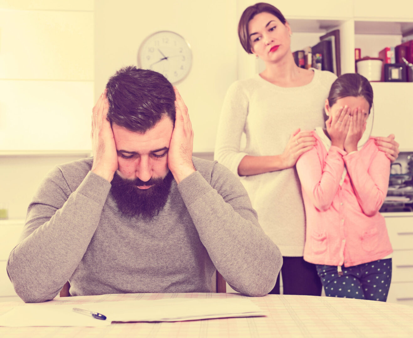 A man sits at a table holding his head, looking at papers. A woman stands behind him with a hand on a child's shoulder. The child covers her face with her hands. A clock is on the wall.