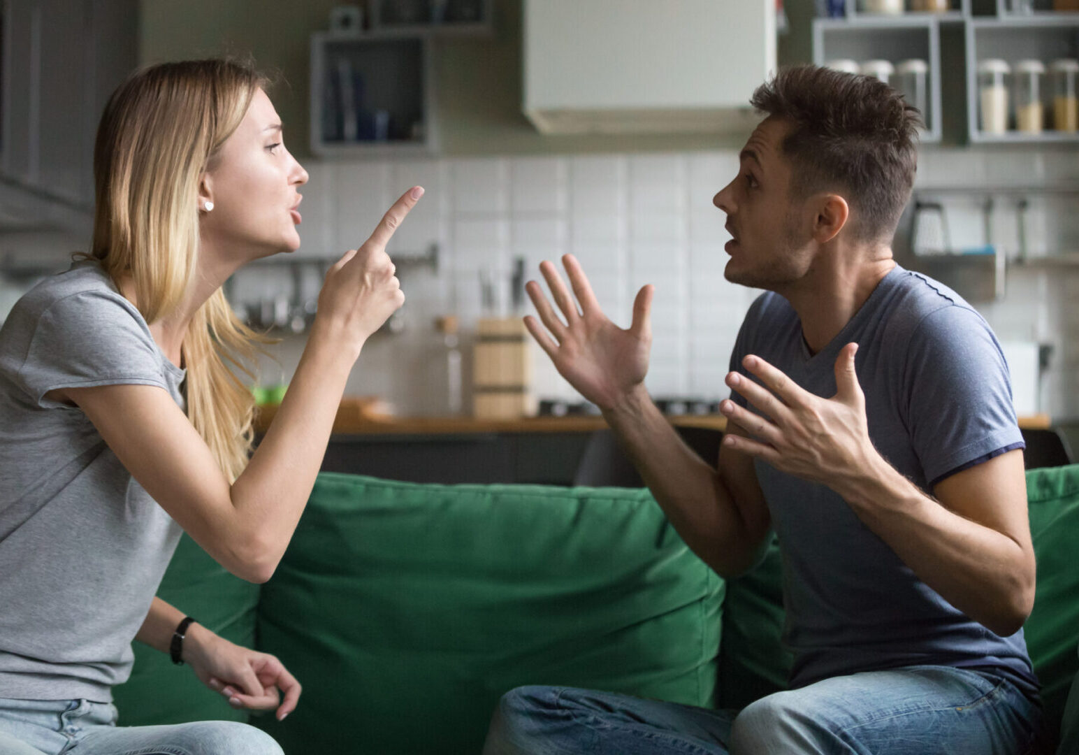 A man and woman are sitting on a green couch in a kitchen, engaged in a heated argument, gesturing with their hands.