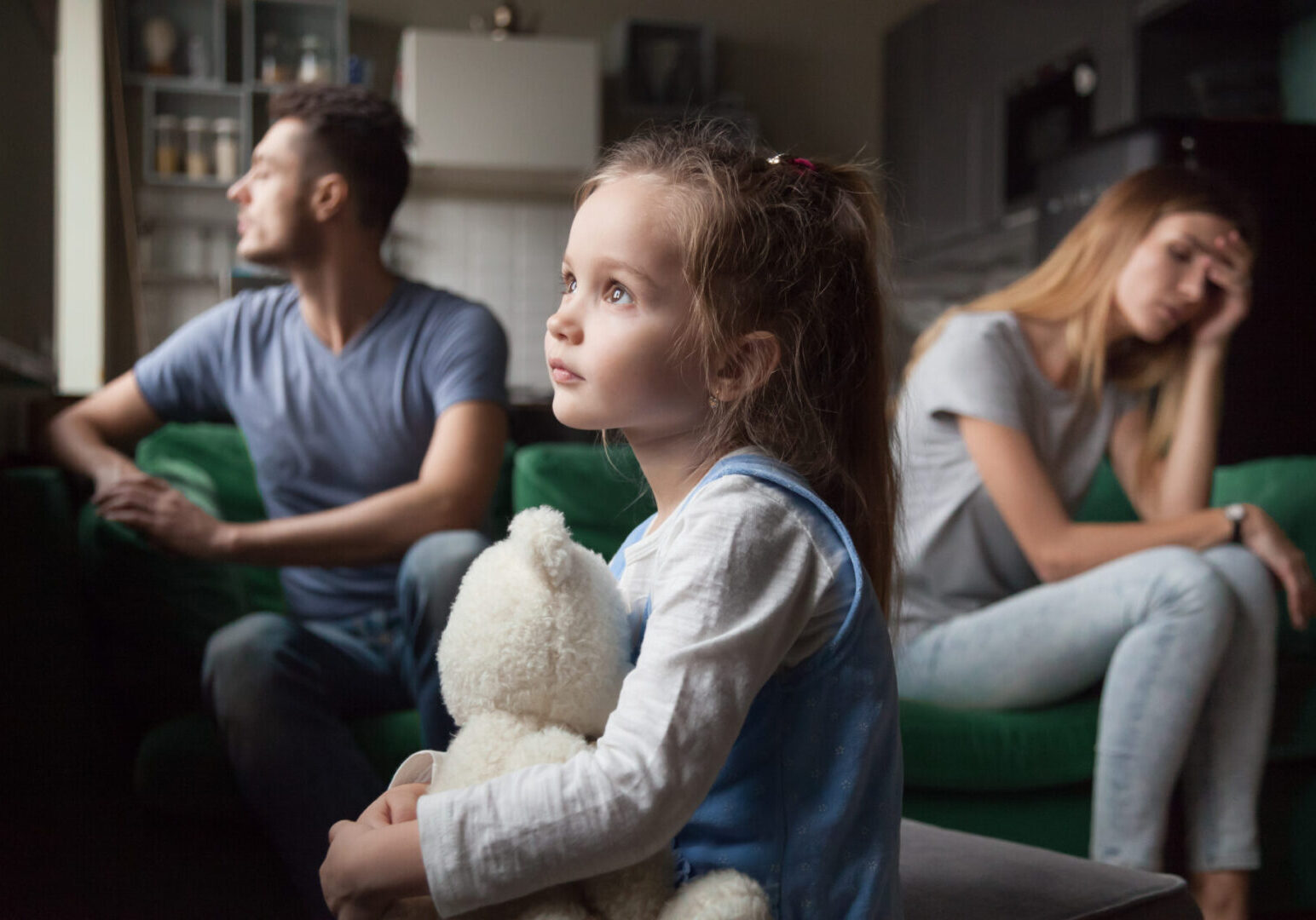 A girl holding a teddy bear looks out the window, while a man and woman sit on a green couch in the background, appearing deep in thought or concern.