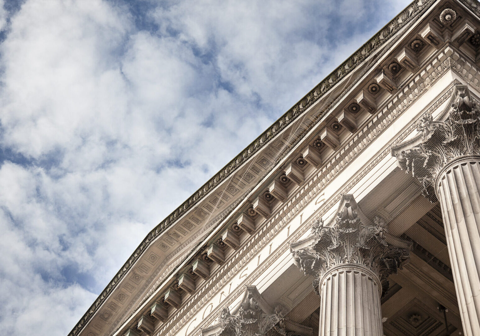 Low-angle view of a neoclassical building facade with ornate Corinthian columns against a partly cloudy sky.